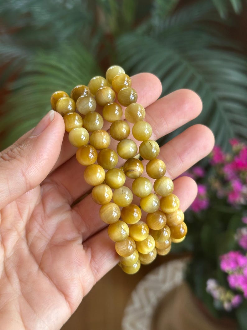 May include: A set of three golden yellow beaded bracelets. The round beads have a polished finish and are strung together. The bracelets are held in a hand, with a blurred green and pink floral background.