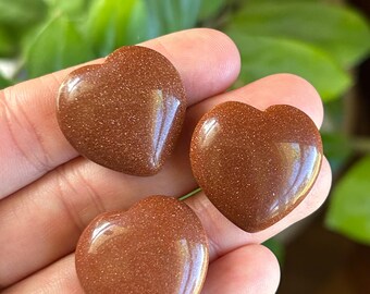 May include: Three polished heart-shaped stones made of brown goldstone, featuring a smooth, glossy surface with embedded metallic glitter particles that catch the light. Each stone is shaped into a symmetrical heart with a rounded top and pointed base.