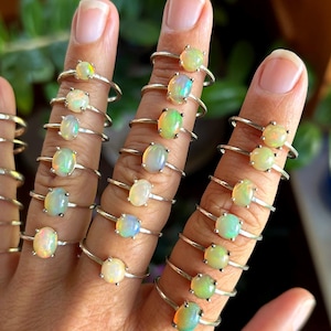 May include: A collection of thin silver rings, each featuring a single oval-shaped opal gemstone. The opals display iridescent flashes of color, including white, green, orange, and blue, set in simple metal bands worn across multiple fingers.
