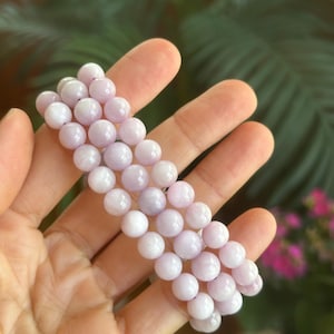 K&ouml;nnte beinhalten: Ein helllila Perlenarmband. Das Armband besteht aus runden, polierten Perlen in verschiedenen Lavendel- und Wei&szlig;t&ouml;nen. Die Perlen sind zu einem flexiblen Band aufgereiht. Der Hintergrund ist verschwommen, mit gr&uuml;nem Laub und rosa Bl&uuml;ten.