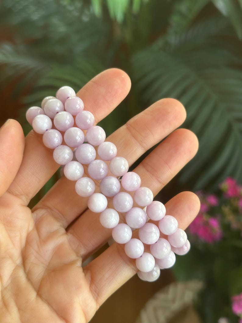 K&ouml;nnte beinhalten: Ein helllila Perlenarmband. Das Armband besteht aus runden, polierten Perlen in verschiedenen Lavendel- und Wei&szlig;t&ouml;nen. Die Perlen sind zu einem flexiblen Band aufgereiht. Der Hintergrund ist verschwommen, mit gr&uuml;nem Laub und rosa Bl&uuml;ten.