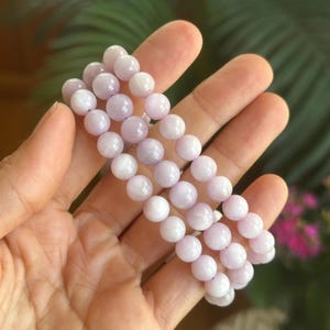 K&ouml;nnte beinhalten: Drei Armb&auml;nder aus hellvioletten, runden Perlen werden in einer Hand gehalten. Die Perlen haben ein glattes, poliertes Aussehen. Der Hintergrund ist verschwommen, mit gr&uuml;nem Laub und rosa Bl&uuml;ten.