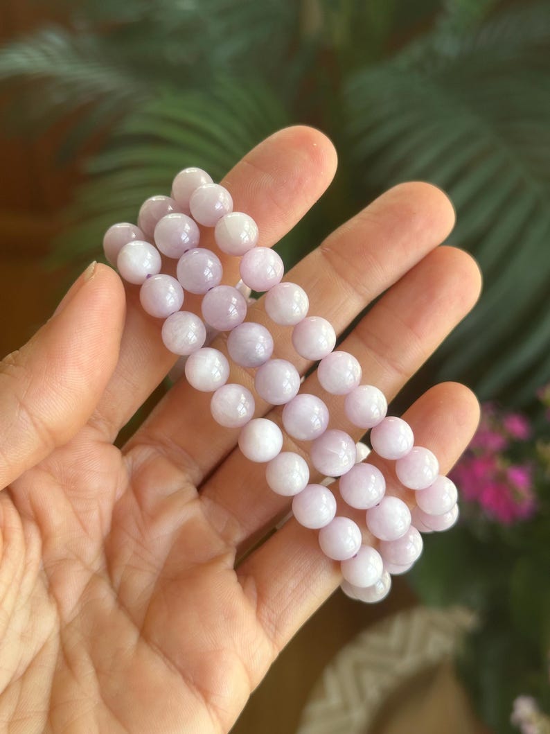 K&ouml;nnte beinhalten: Drei Armb&auml;nder aus hellvioletten, runden Perlen werden in einer Hand gehalten. Die Perlen haben ein glattes, poliertes Aussehen. Der Hintergrund ist verschwommen, mit gr&uuml;nem Laub und rosa Bl&uuml;ten.