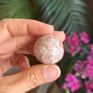 May include: A spherical, polished stone featuring a swirling pattern of pink, white, and grey. The stone is held in a hand, with a blurred backdrop of green leaves and pink flowers.