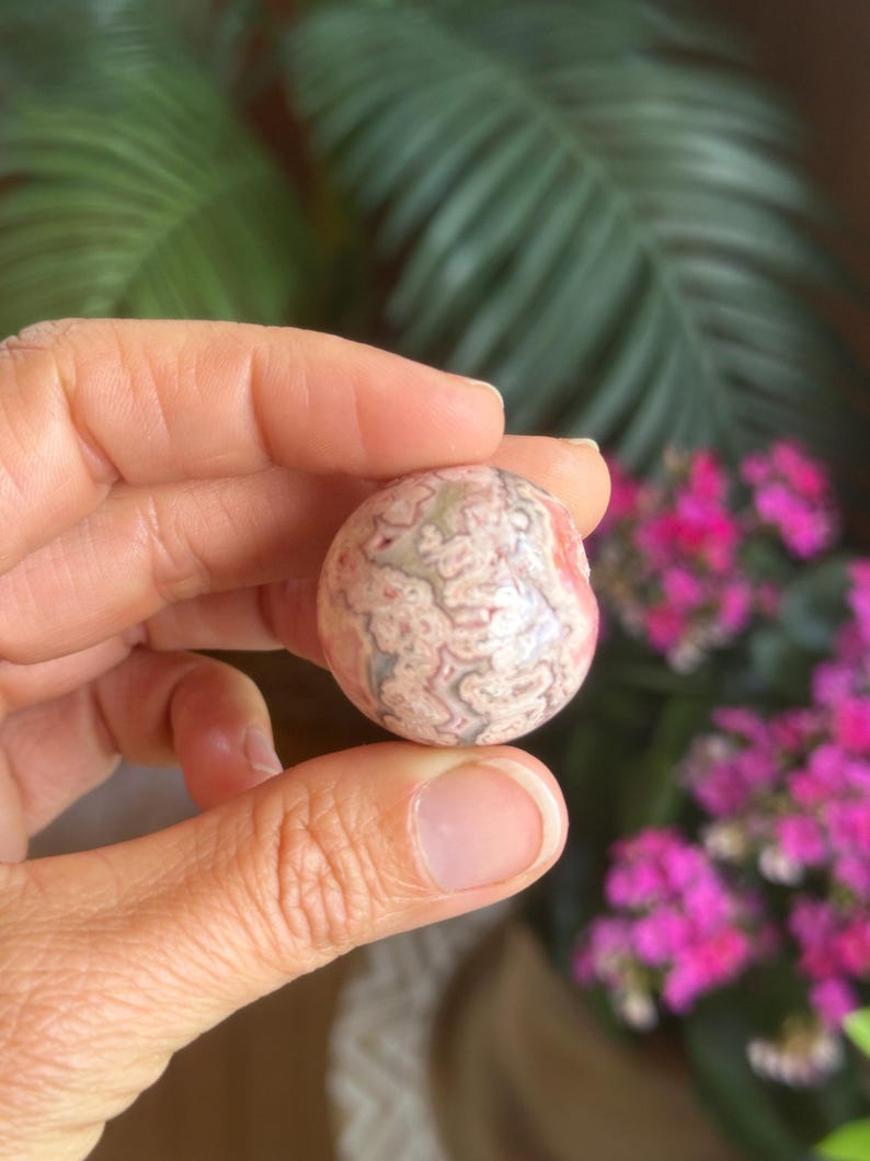 May include: A spherical, polished stone featuring a swirling pattern of pink, white, and grey. The stone is held in a hand, with a blurred backdrop of green leaves and pink flowers.