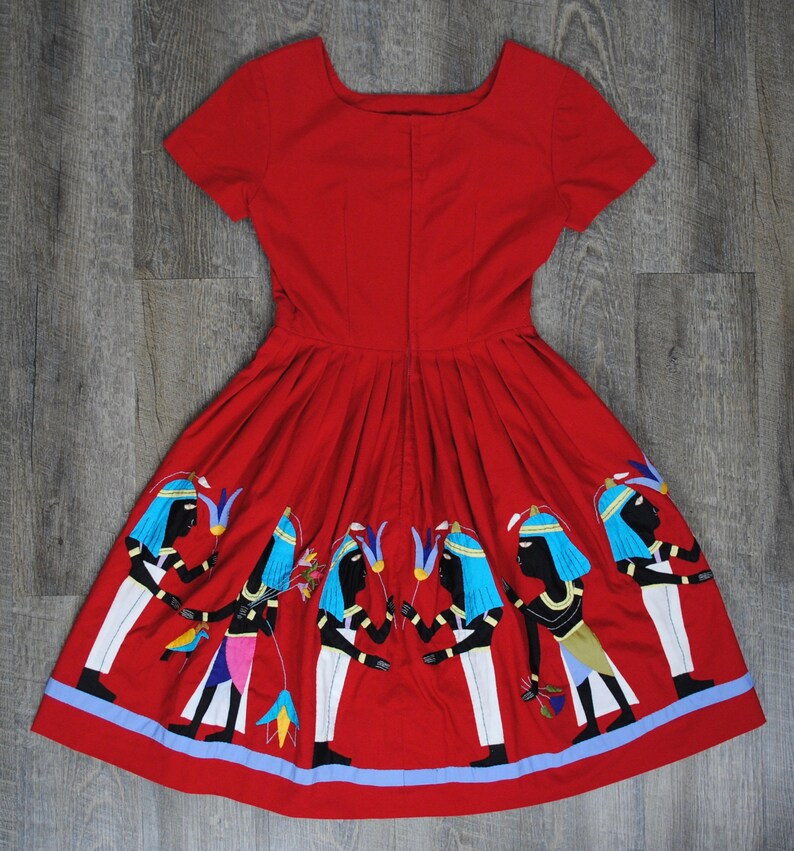May include: A red dress with a scoop neck and short sleeves. The dress has a pleated skirt with embroidered figures of people in blue and white.