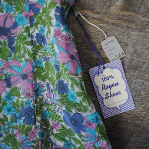 May include: A white dress with a floral print in shades of pink, blue, and green. The dress is made of 100% rayon sheer fabric. The tag says "100% Rayon Sheer" and "Exclusive of Ornamentation".