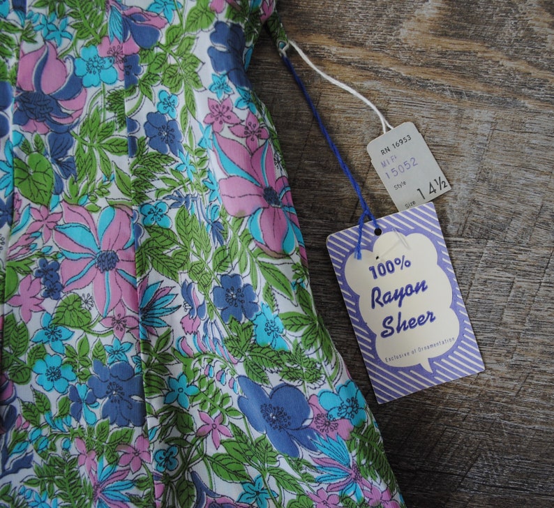 May include: A white dress with a floral print in shades of pink, blue, and green. The dress is made of 100% rayon sheer fabric. The tag says "100% Rayon Sheer" and "Exclusive of Ornamentation".