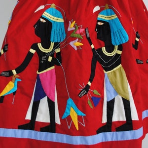 May include: Red cotton skirt with embroidered figures of Egyptian pharaohs in black, blue, yellow, and white. The skirt has a blue trim.