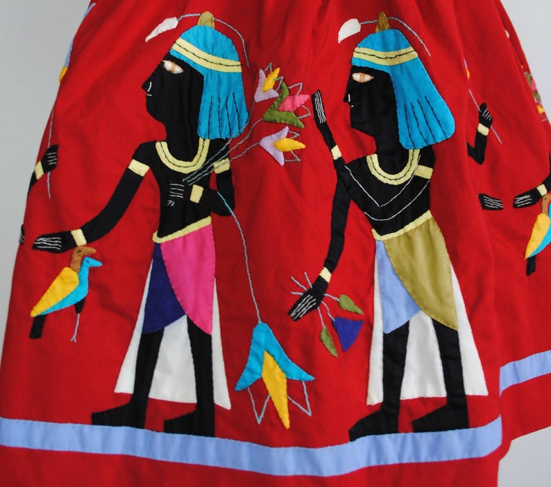 May include: Red cotton skirt with embroidered figures of Egyptian pharaohs in black, blue, yellow, and white. The skirt has a blue trim.