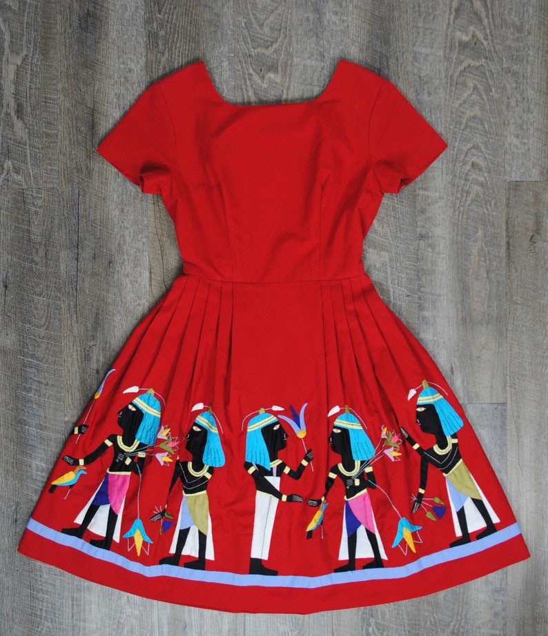 May include: A red dress with a square neckline and short sleeves. The skirt of the dress is decorated with an embroidered design of figures in blue, white, and black.
