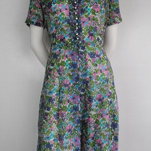 May include: A vintage floral print dress with a short sleeve, a v-neck, and a button-up front. The dress is made of a light-weight fabric and has a white background with pink, blue, and green flowers.