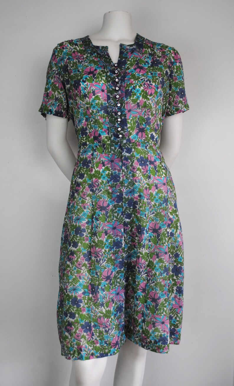 May include: A vintage floral print dress with a short sleeve, a v-neck, and a button-up front. The dress is made of a light-weight fabric and has a white background with pink, blue, and green flowers.