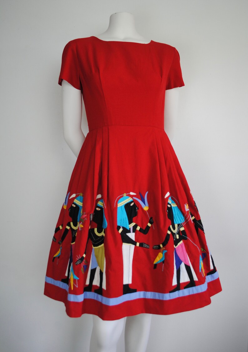 May include: A red vintage dress with a fitted bodice and a full skirt. The skirt features an embroidered design of figures in a procession, with blue, yellow, and white accents.