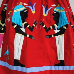 May include: Red cotton skirt with embroidered figures of Egyptian deities holding flowers. The skirt has a blue trim.