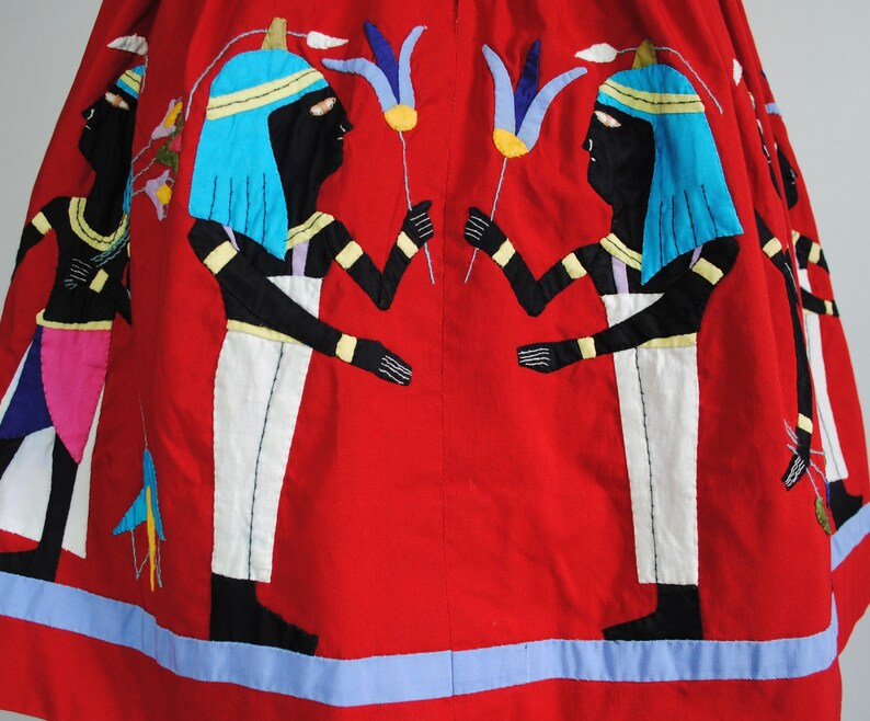 May include: Red cotton skirt with embroidered figures of Egyptian deities holding flowers. The skirt has a blue trim.
