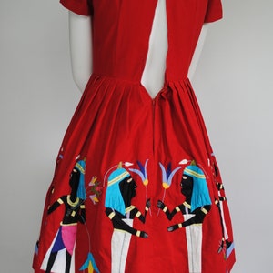 May include: A red dress with a keyhole back and embroidered figures in blue, white, and black. The dress has a full skirt and a blue trim.