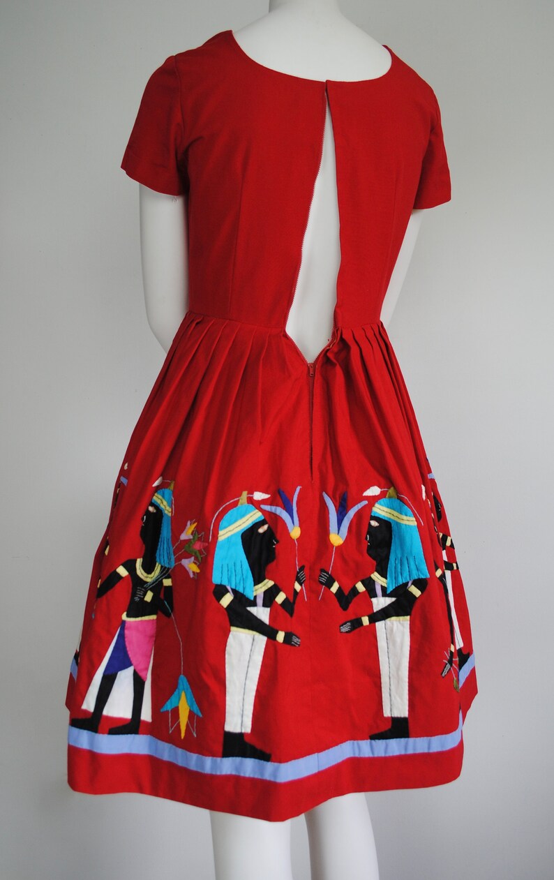 May include: A red dress with a keyhole back and embroidered figures in blue, white, and black. The dress has a full skirt and a blue trim.