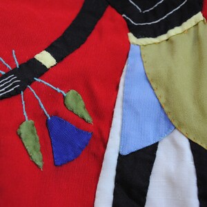 May include: A red fabric background with a black, white, blue, and green applique design. The design features a stylized figure with a long, flowing skirt and a hand holding a bouquet of flowers.
