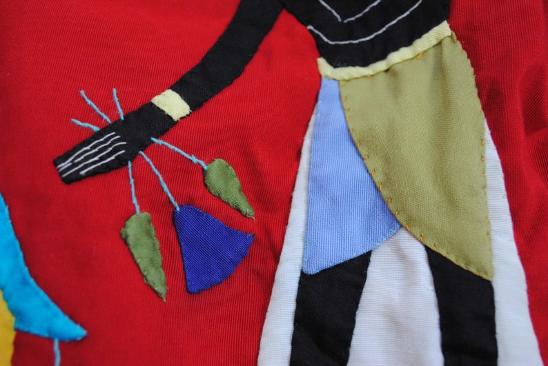 May include: A red fabric background with a black, white, blue, and green applique design. The design features a stylized figure with a long, flowing skirt and a hand holding a bouquet of flowers.