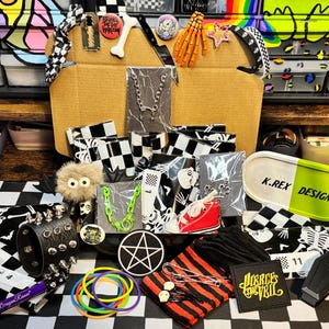 May include: A cardboard box filled with assorted accessories including a black spiked wristband, a pentagram patch, a neon green chain, a red sneaker keychain, skeleton-patterned fabric, hair clips, a K.REX DESIGNS tray, and various pins.