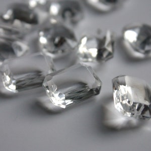 May include: A group of clear, faceted glass crystals, some with a rectangular shape, scattered on a white surface.