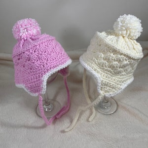 May include: Two crocheted baby hats with pom poms. One hat is pink with a white trim and the other is white with a white trim.