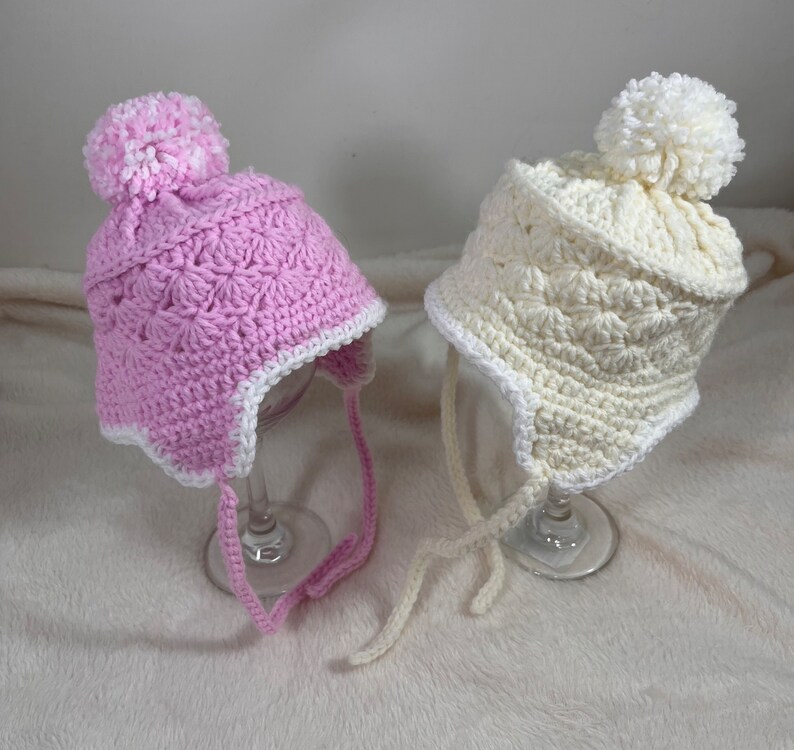 May include: Two crocheted baby hats with pom poms. One hat is pink with a white trim and the other is white with a white trim.