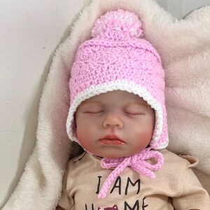 May include: A pink crocheted baby hat with a white pom-pom on top and a white crocheted brim. The hat has ear flaps and a tie under the chin.