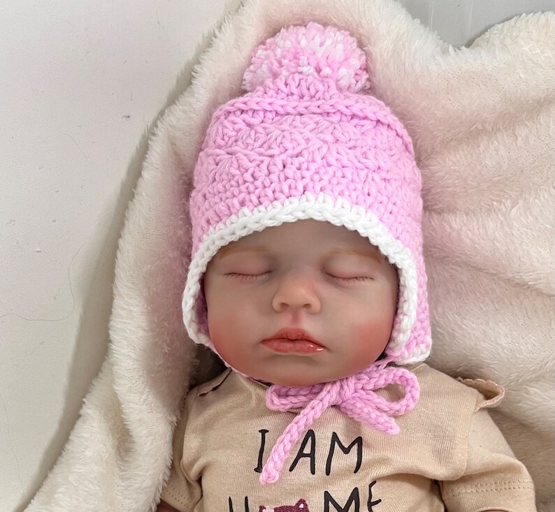 May include: A pink crocheted baby hat with a white pom-pom on top and a white crocheted brim. The hat has ear flaps and a tie under the chin.