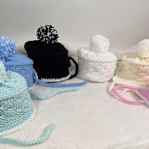 May include: Five crocheted hats with pom-poms in various colors, including blue, white, pink, and black. The hats have a crocheted brim and a tie.