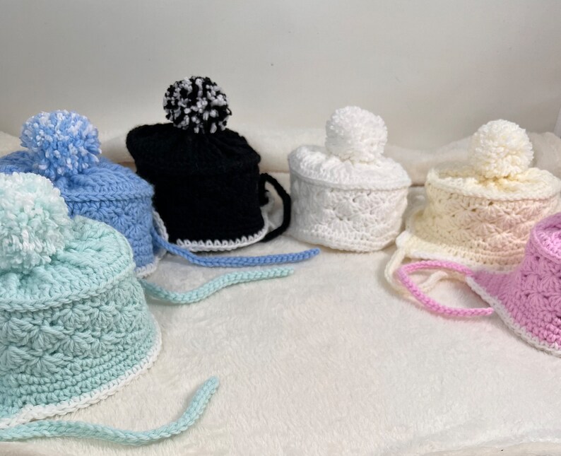 May include: Five crocheted hats with pom-poms in various colors, including blue, white, pink, and black. The hats have a crocheted brim and a tie.