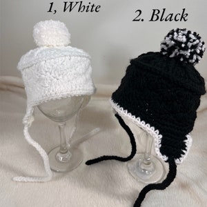 May include: Two crocheted hats with pom-poms. One hat is white with a white pom-pom and the other is black with a black and white pom-pom. Both hats have white trim and long ties.
