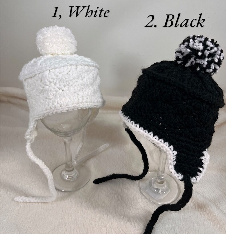 May include: Two crocheted hats with pom-poms. One hat is white with a white pom-pom and the other is black with a black and white pom-pom. Both hats have white trim and long ties.