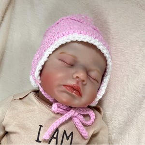 May include: A sleeping baby doll wearing a pink and white crocheted hat with a bow. The doll is wearing a tan shirt with the text "I AM" printed on it.