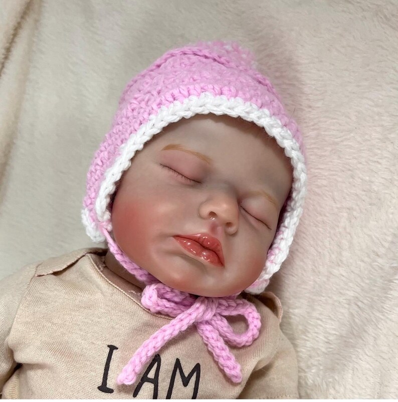 May include: A sleeping baby doll wearing a pink and white crocheted hat with a bow. The doll is wearing a tan shirt with the text "I AM" printed on it.