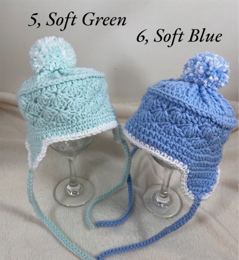 May include: Two crocheted hats with white pom-poms. The hat on the left is light blue and the hat on the right is light green. Both hats have white trim around the brim and are tied with long strings.