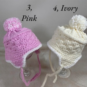 May include: Two crocheted baby hats with pom poms. One hat is pink with a white trim and the other is ivory with a white trim. The hats have ties.