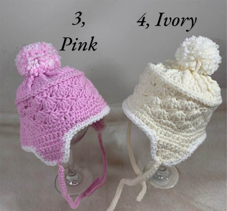 May include: Two crocheted baby hats with pom poms. One hat is pink with a white trim and the other is ivory with a white trim. The hats have ties.