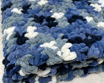 May include: A soft, blue, gray, and white crocheted blanket with a fluffy texture. The blanket is folded in half and has a subtle pattern.