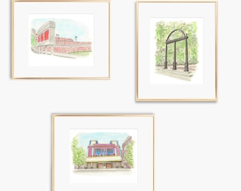 Pode incluir: Três aquarelas emolduradas de marcos icônicos em Atenas, Geórgia. A primeira pintura mostra um estádio com assentos vermelhos e uma multidão de pessoas. A segunda pintura mostra o arco icônico conhecido como 'Arch' na Universidade da Geórgia. A terceira pintura mostra a fachada do Georgia Theatre.