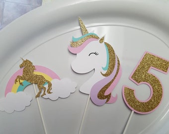 May include: Three cake toppers featuring a unicorn, a rainbow with a cloud, and the number 5. The unicorn is white with pink, purple, and blue glitter accents. The rainbow is yellow, pink, and blue with a white cloud. The number 5 is gold glitter with a pink outline.
