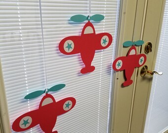 May include: Red paper airplane decorations with teal propellers and white star accents are suspended in front of a window. The airplanes are hanging from thin white strings. The background includes white blinds and a white door.