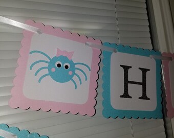 May include: A pink and blue banner with the word "Happy" in silver glitter letters. The banner has a scalloped edge and is strung together with white ribbon. The first banner features a blue spider with a pink bow on its head.