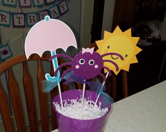 May include: A purple glitter spider, a pink umbrella, and a yellow sun with a smiling face are arranged in a purple bucket filled with white shredded paper. The decorations are on a white table.