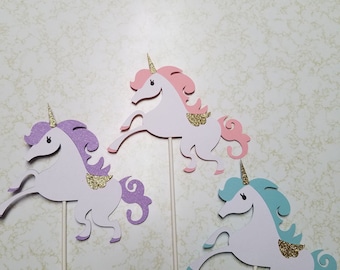 May include: Three whimsical unicorn cutouts in white, purple, pink, and blue with gold glitter accents. Each unicorn is attached to a wooden stick, perfect for cake toppers or party decorations. The unicorns have a smiling face and are in a running pose.