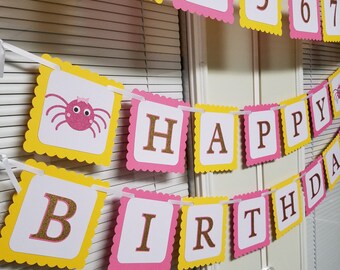 May include: A pink and yellow banner with the words "Happy Birthday" and a pink spider with a bow. The banner is decorated with scalloped edges.
