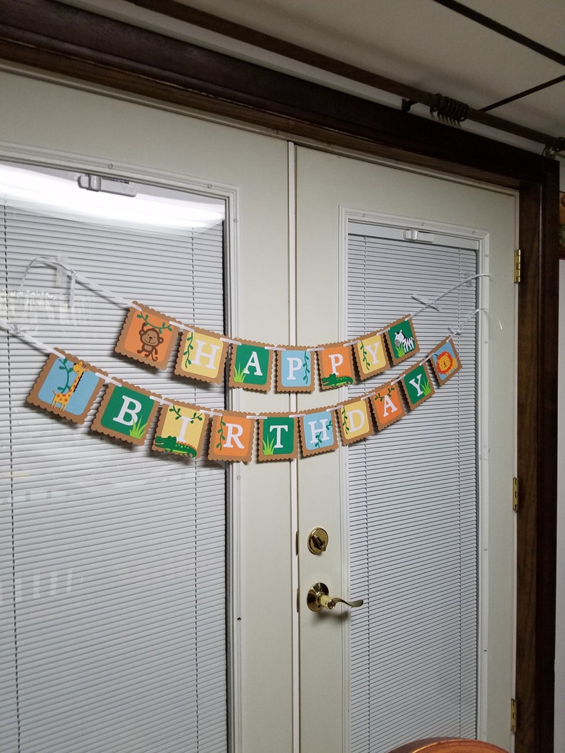 May include: A banner with the words "Happy Birthday" in colorful letters with jungle animal illustrations. The banner is strung across two white doors with blinds.