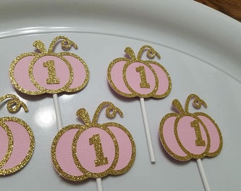 May include: Four pink and gold glitter pumpkin cupcake toppers with the number one on each. The pumpkins are layered and have a gold glitter swirl on top.
