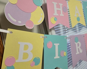 May include: A pastel-colored banner with the words "Happy Birthday" in white letters. The banner is decorated with pink, yellow, and blue circles.